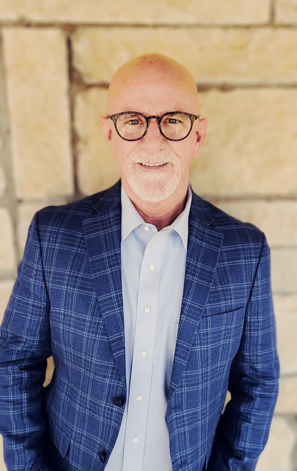 Bald man with glasses and goatee wearing a blue plaid blazer against a stone wall.
